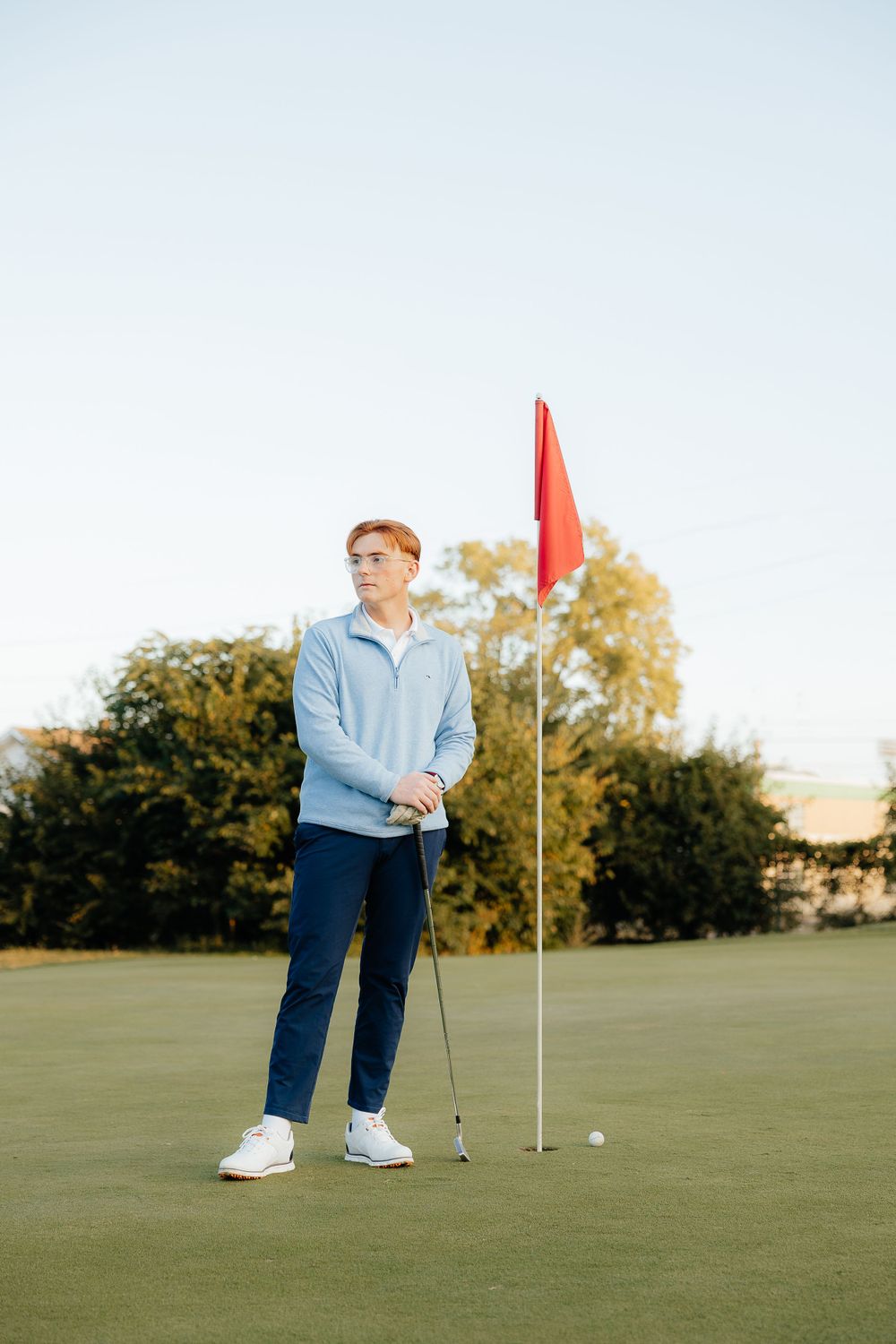 Golfer stands near red flag on putting green during autumn afternoon.