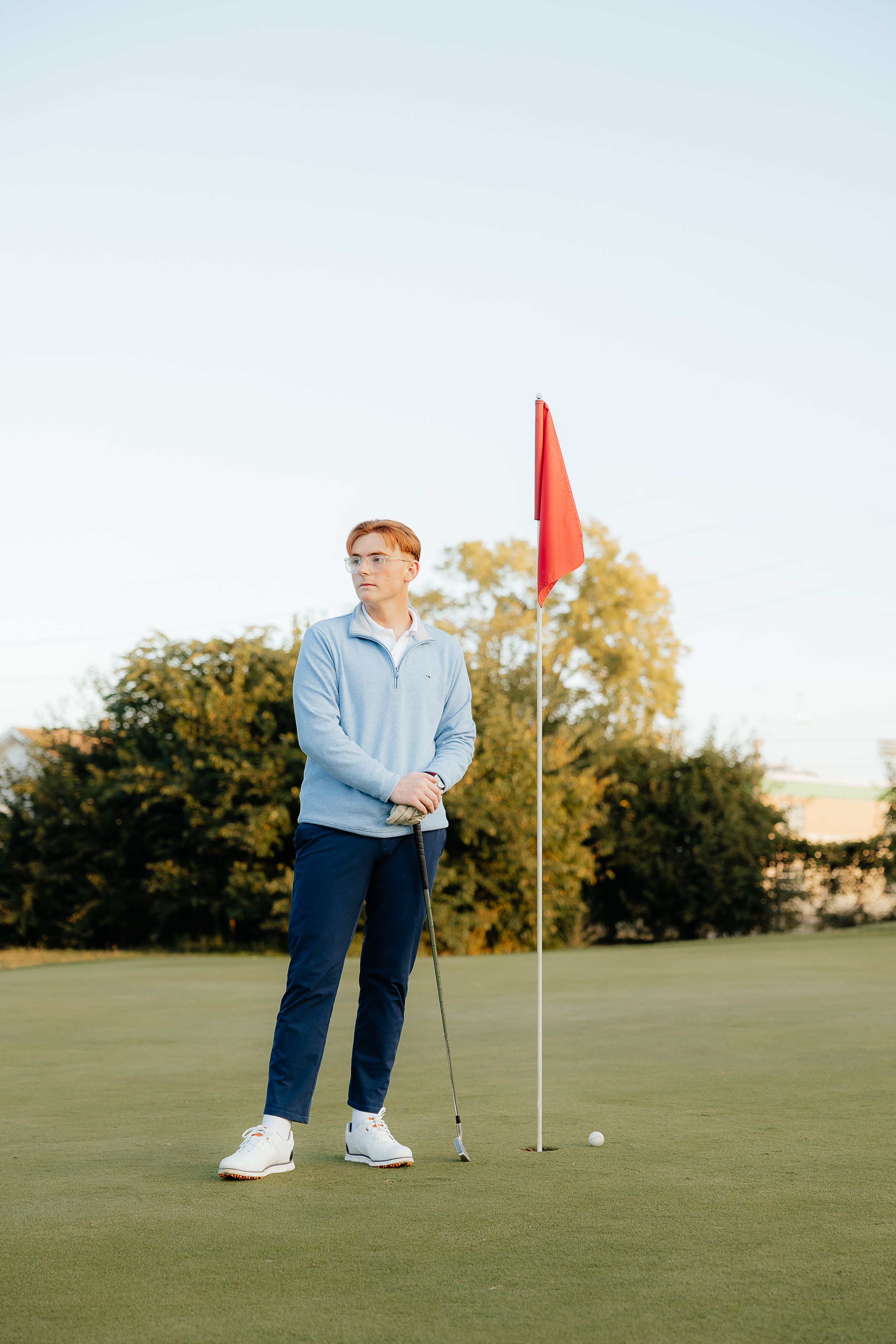 Golfer stands near red flag on putting green during autumn afternoon.