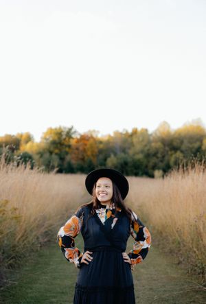 Portrait in tall grass wearing black dress and wide-brimmed hat with autumn foliage behind.