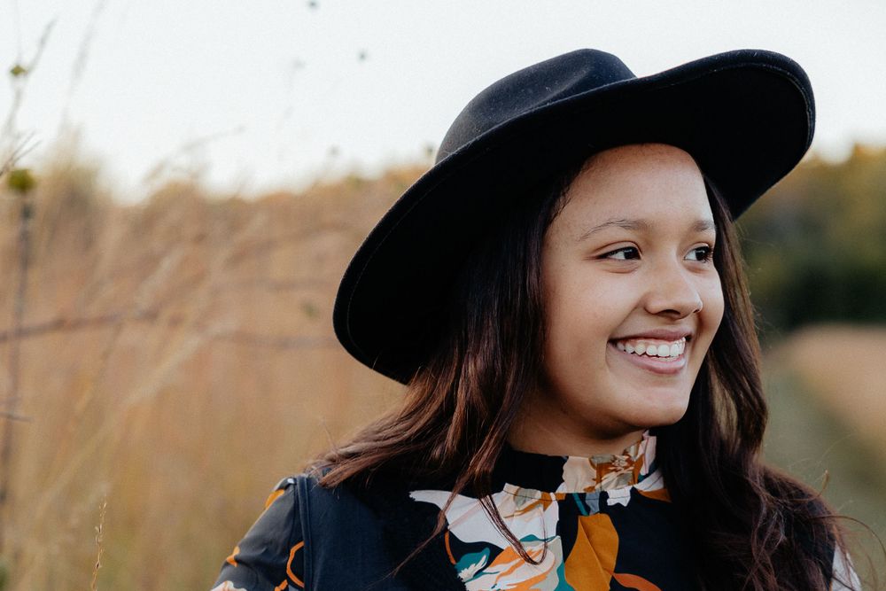 A person wearing a black hat and floral dress poses in a field during golden hour.