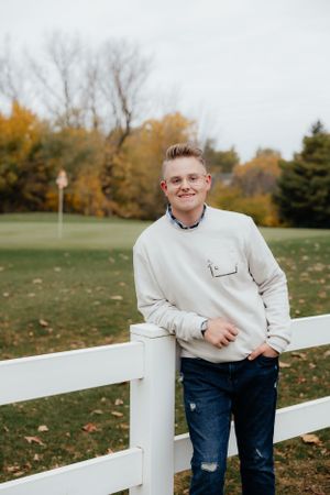 Casual outdoor portrait by white fence with fall colors and trees in background.