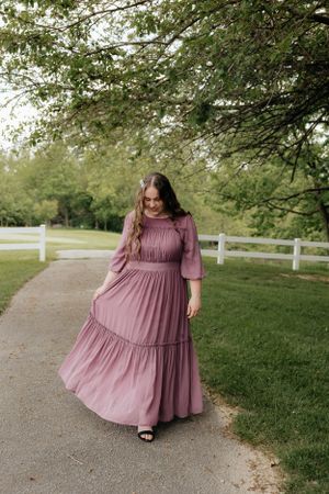 A flowing mauve dress with gathered details shown twirling on a tree-lined path.
