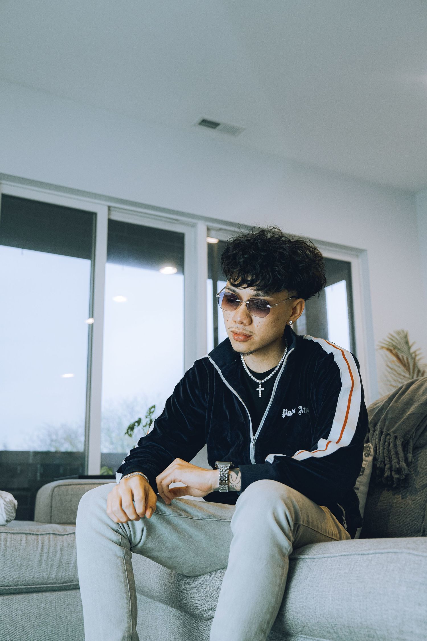 Casual portrait of someone wearing sunglasses and tracksuit sitting on grey couch.