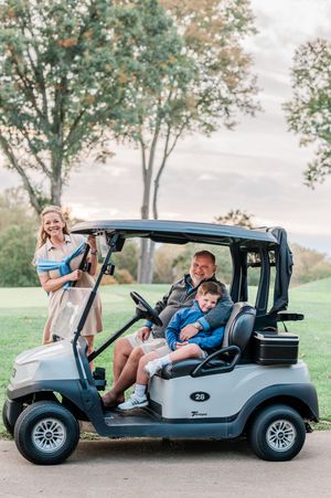 Three people enjoying a sunny day riding in a gray golf cart on a scenic course with autumn trees in background.