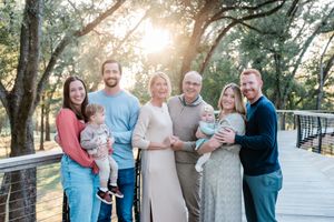 Multi-generational family portrait outdoors on a wooden bridge, smiling in golden sunset light among trees.
