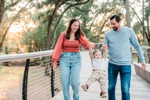 Happy family with baby walking on a wooden bridge outdoors, surrounded by lush green trees in warm golden light.