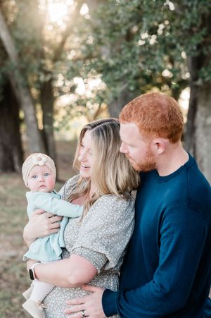 A young family with a redheaded father and blonde mother smiling at their baby girl outdoors among trees in golden light.