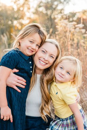 A smiling woman with two young blonde children hugging outdoors in a golden sunlit field.