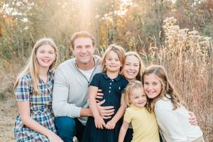A smiling family of six poses outdoors in a sunny autumn field, with a father and five children hugging closely.