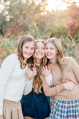 Three smiling teenage girls in cozy sweaters embrace outdoors during golden hour fall photography session.