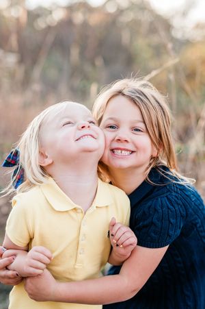Two young siblings hugging outdoors, older girl smiling brightly while younger child looks up, warm natural background.