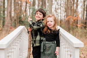 Two young children smiling on a white bridge surrounded by autumn trees, boy holding a stick.