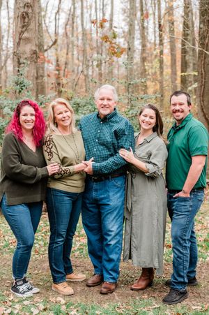 Family of five posing together outdoors in autumn woods, smiling, wearing green and earth tones.