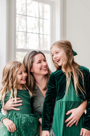 A smiling mother in green embraces her two laughing daughters in matching emerald dresses near a bright window.