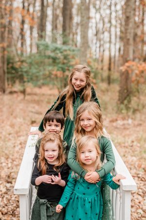 Five children pose together on a white bridge surrounded by autumn foliage in a wooded outdoor setting.