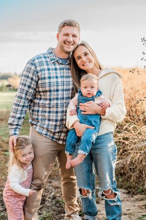 Happy young family of four poses outdoors in fall, parents smiling with toddler girl and baby in denim overalls.