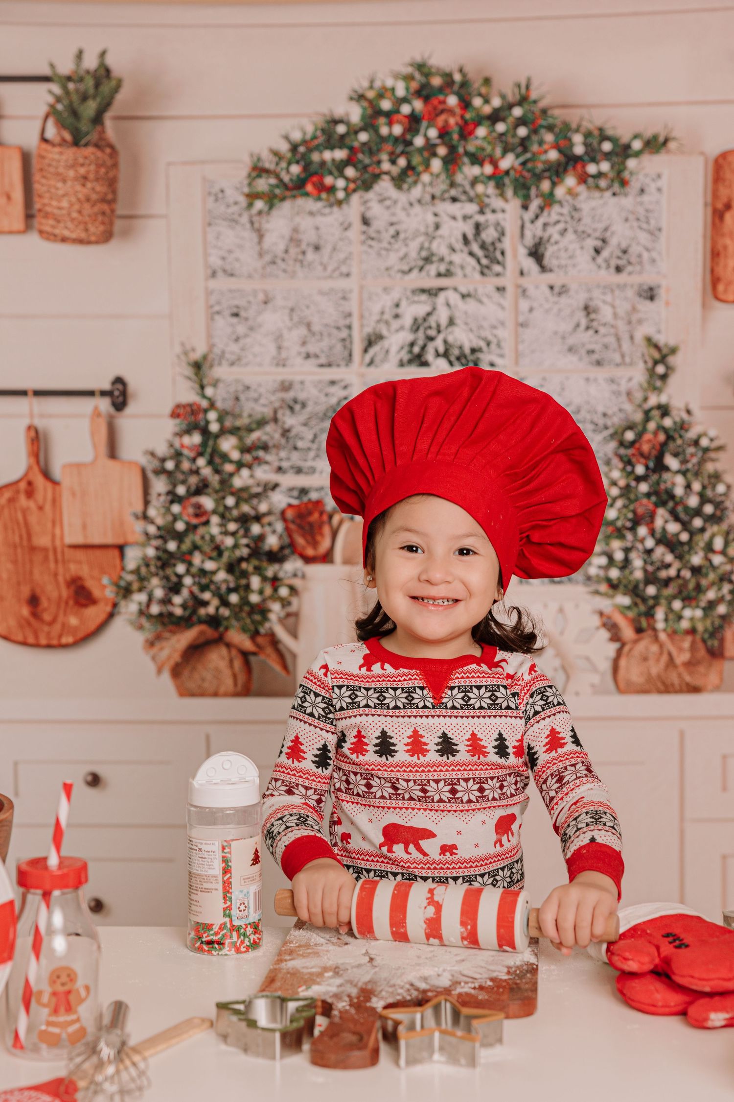 A small baker wearing a red chef hat and holiday sweater makes Christmas cookies at a decorated counter.