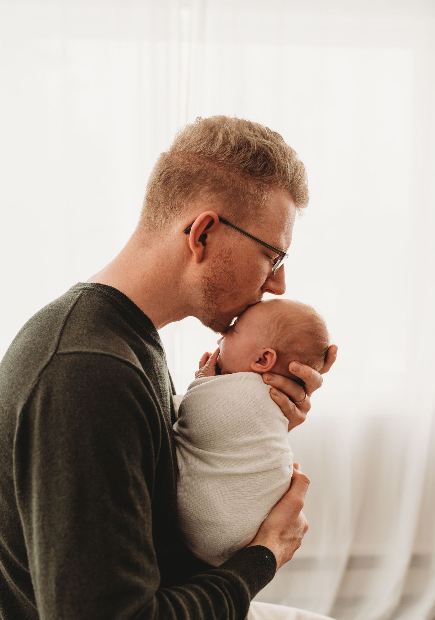 Parent in olive sweater gently holding and kissing newborn baby against white background.