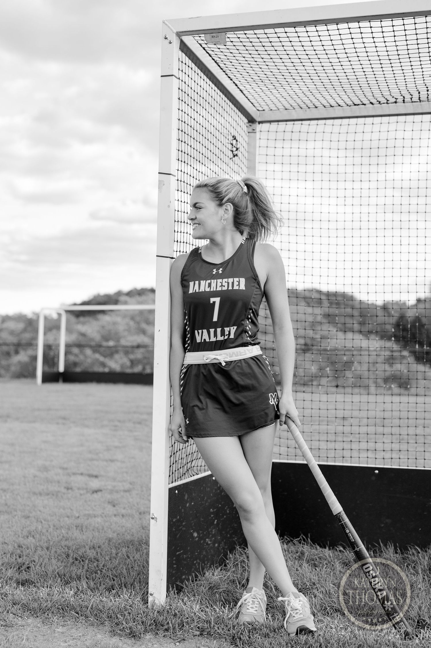 Black and white portrait of athlete leaning against field hockey goal post in team uniform.