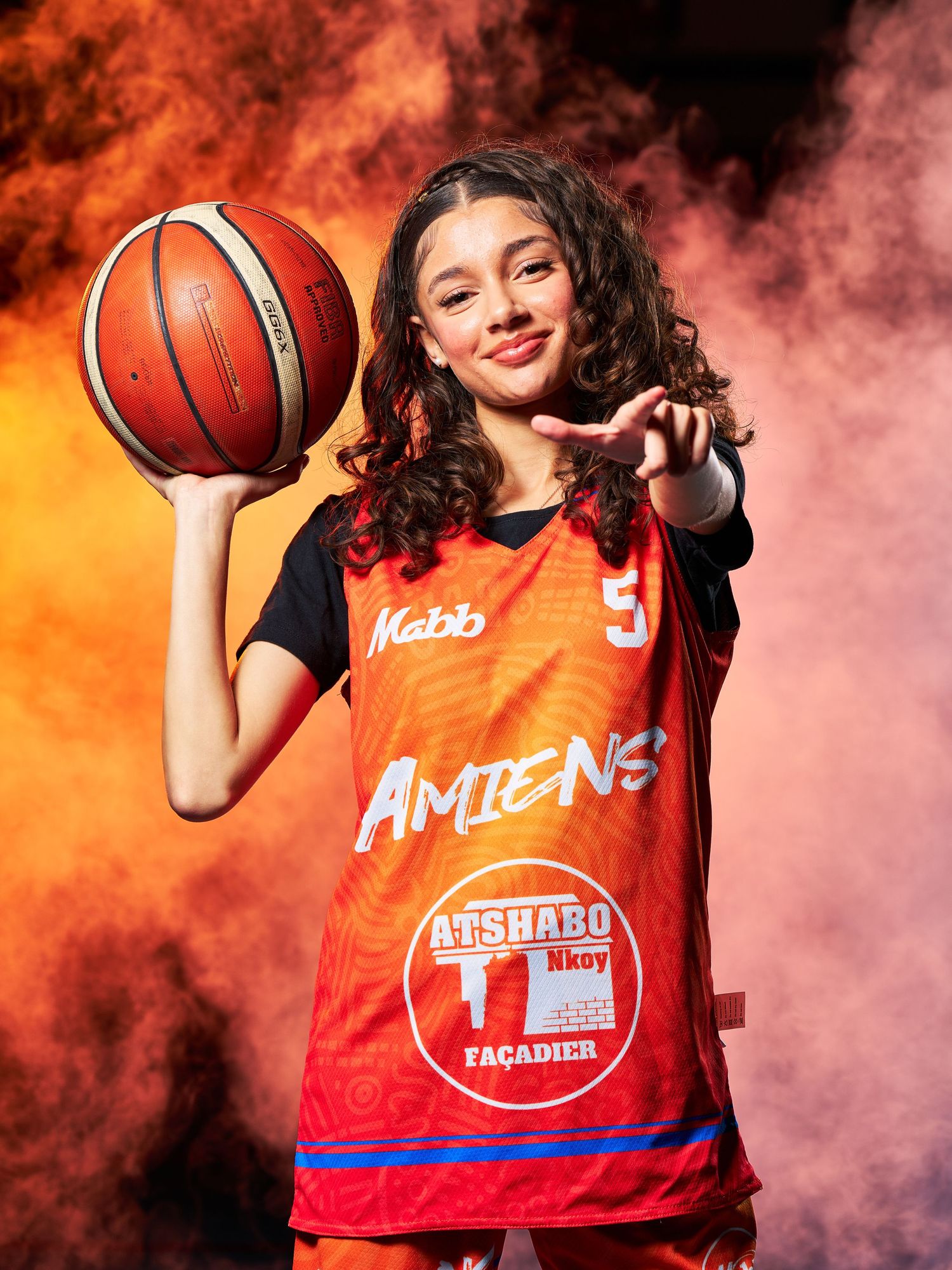 Joueur de basket-ball en maillot rouge d'Amiens pose avec le ballon sur un fond de fumée orange et noir spectaculaire.