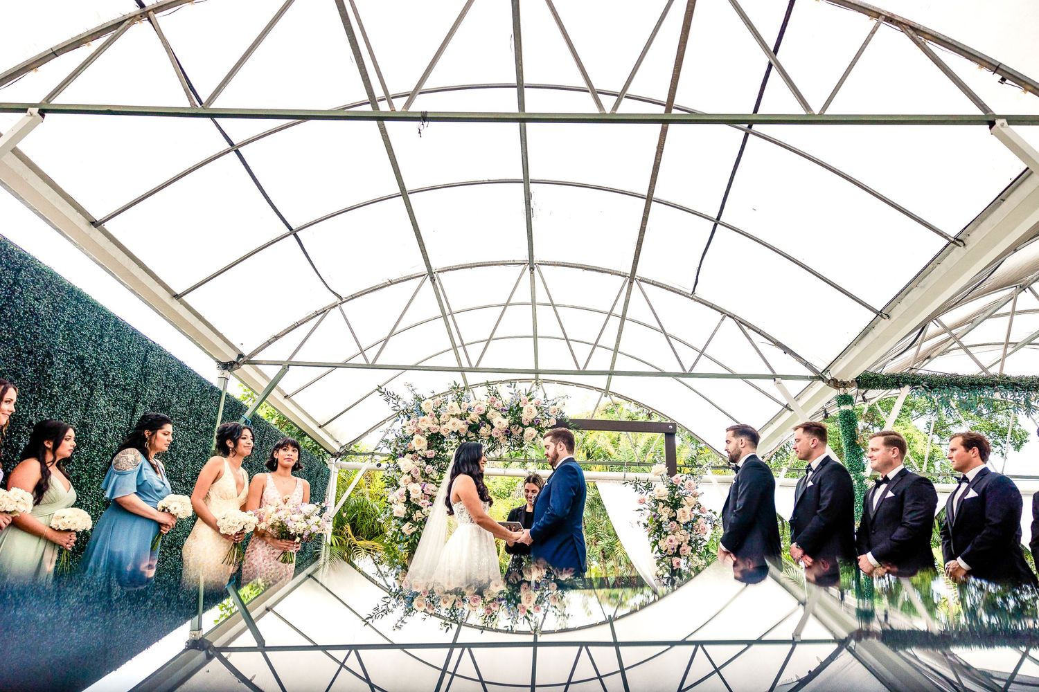 Wedding ceremony taking place under a modern greenhouse structure with arched transparent roof and lush greenery backdrop.