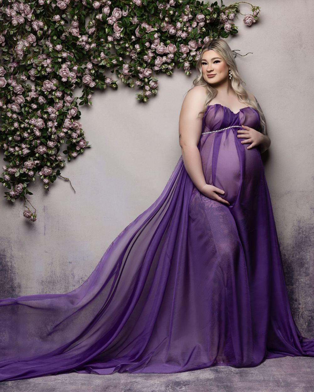 A flowing purple maternity gown and sheer cape create a dramatic silhouette against a floral backdrop.