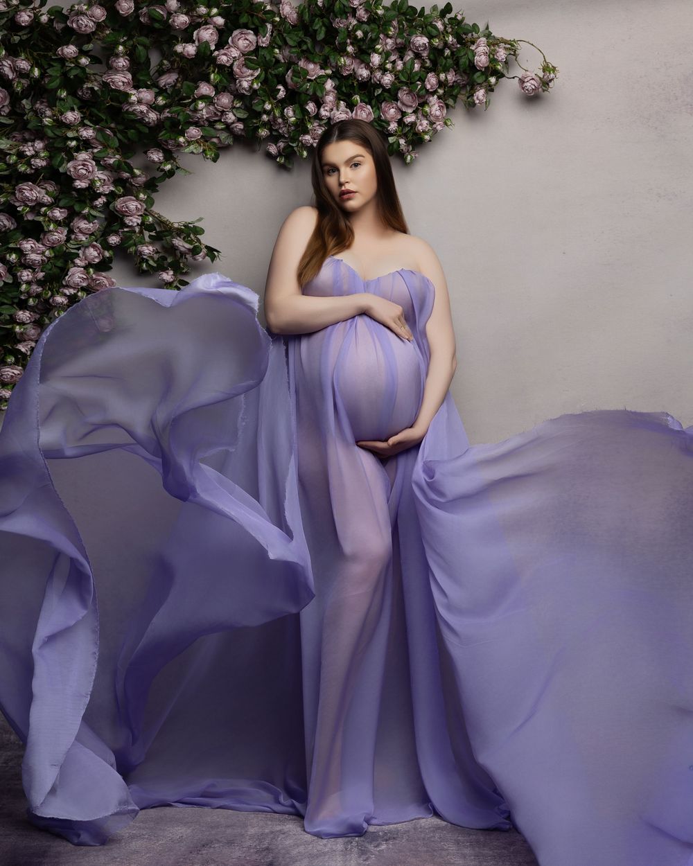 Ethereal maternity photoshoot featuring flowing lavender chiffon gown against pink flowering vine backdrop.