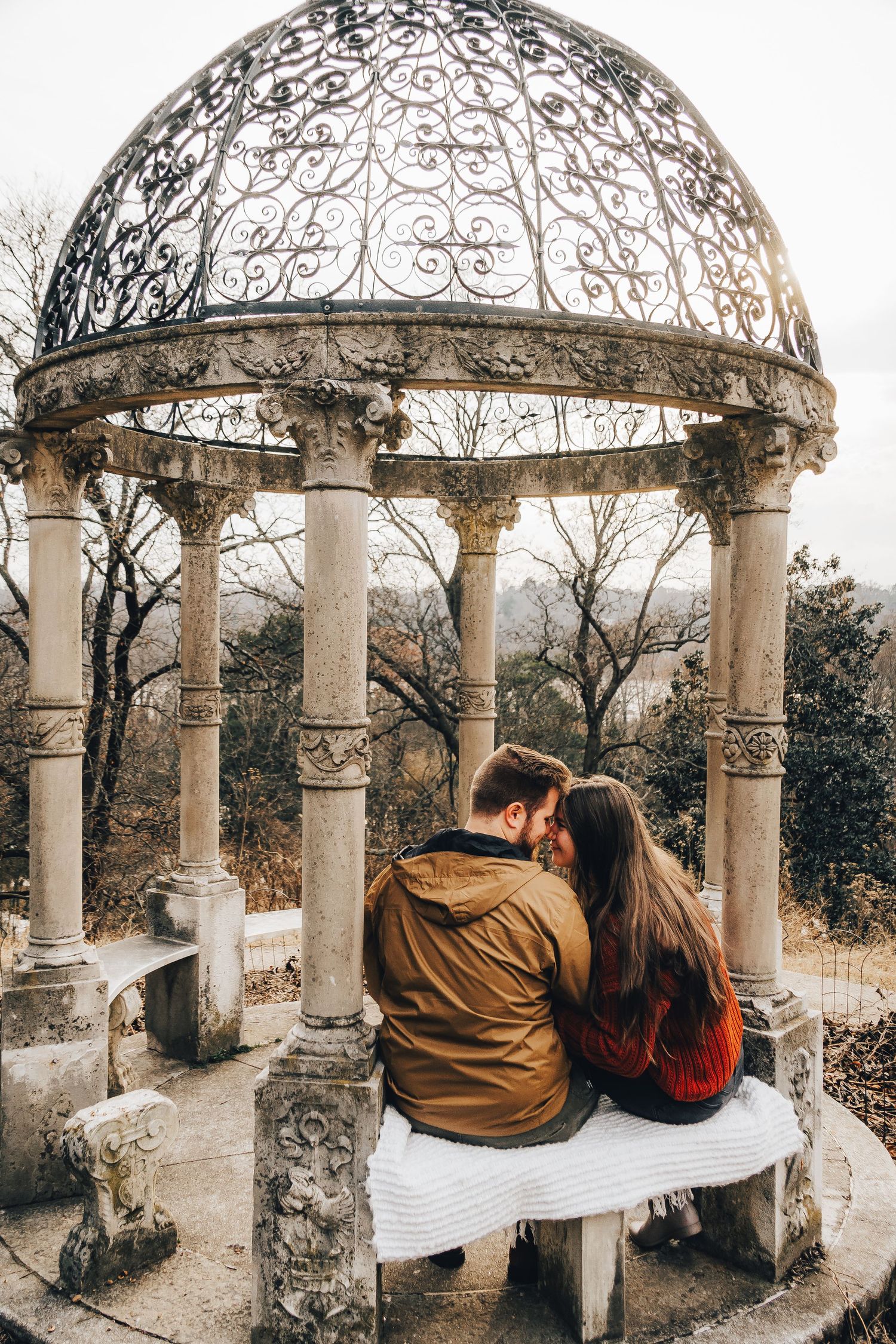 Couple sits close together under ornate stone gazebo in romantic setting.