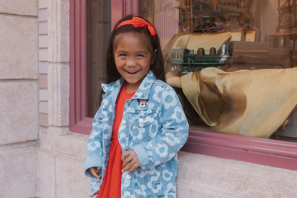Child wearing blue floral jacket and red bow poses by storefront window.