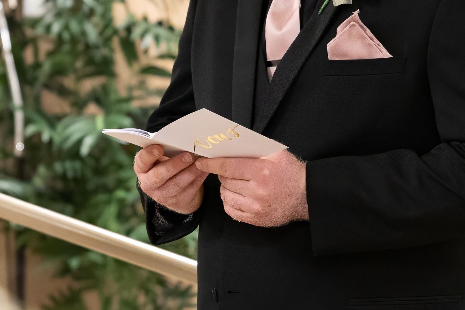 Close-up view of hands holding a card or note while wearing a formal black suit with pink pocket square.