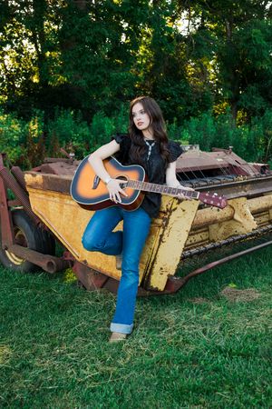 Acoustic guitar player sits on rustic yellow farm equipment in grassy outdoor setting during golden hour.