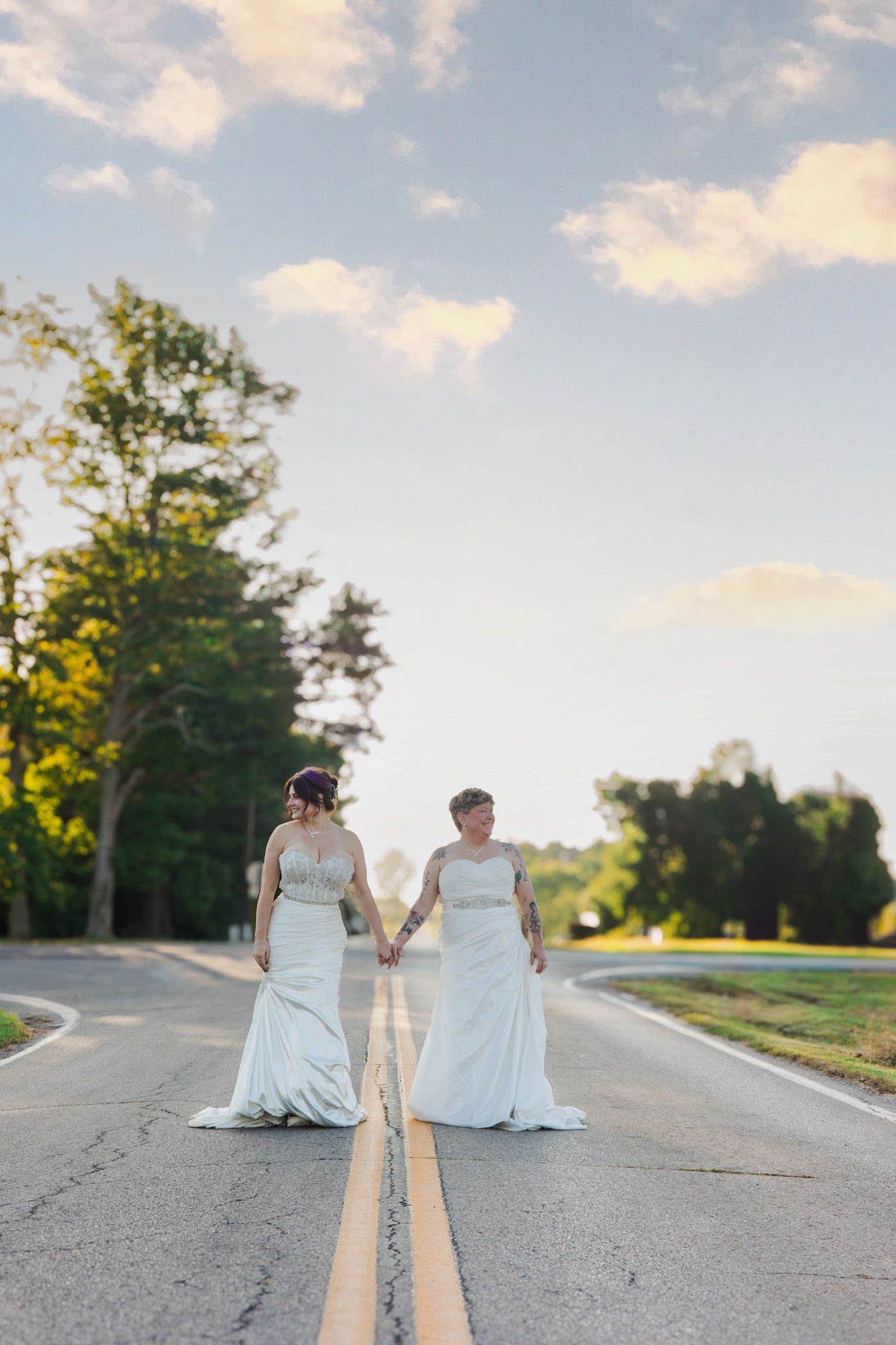Two people in white wedding dresses sit together on an empty country road at sunset.