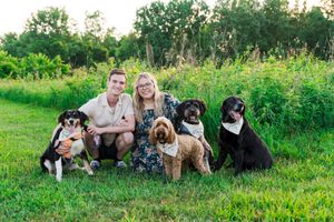 A happy couple sitting on grass with five dogs wearing matching bandanas.