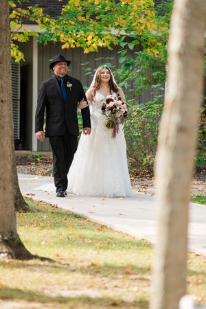 Wedding couple walks along tree-lined path during autumn outdoor ceremony in romantic woodland setting.