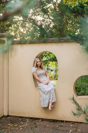 A woman in a polka dot dress sits gracefully on a beige wall in a garden setting during golden hour.