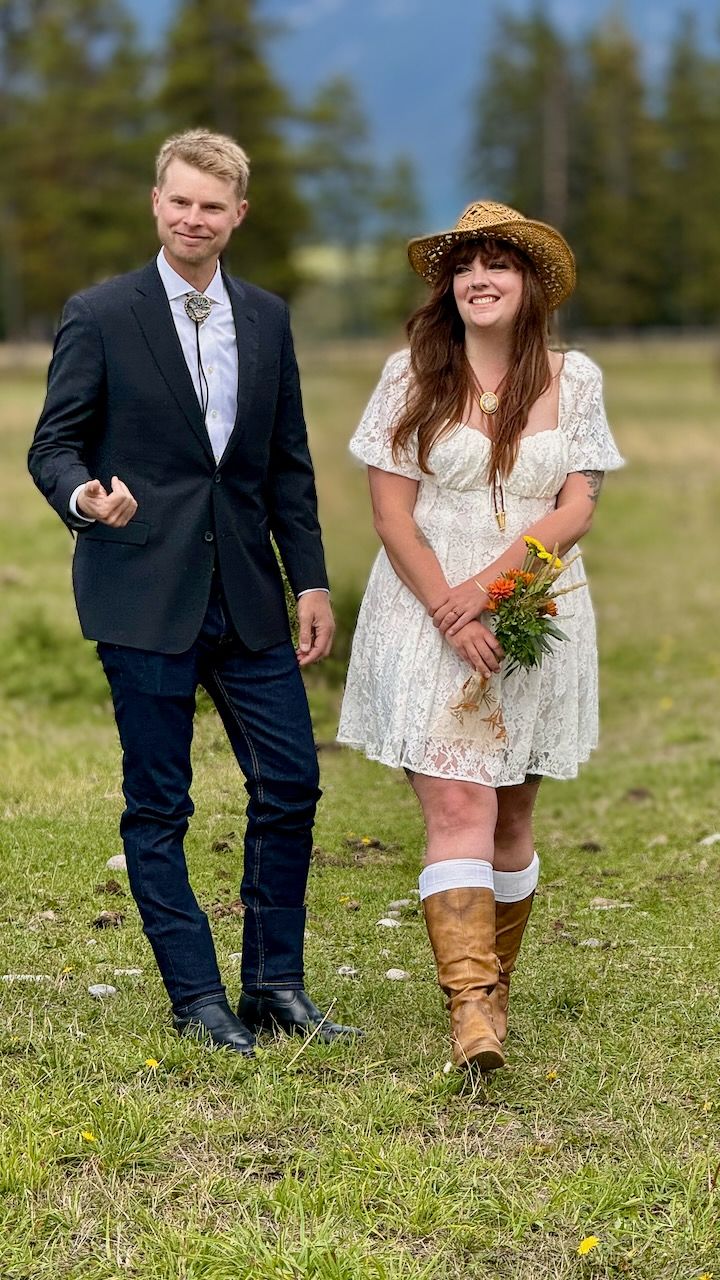 A couple stands in a grassy field - the man in a suit and the woman in a white lace dress with cowboy boots and sun hat.