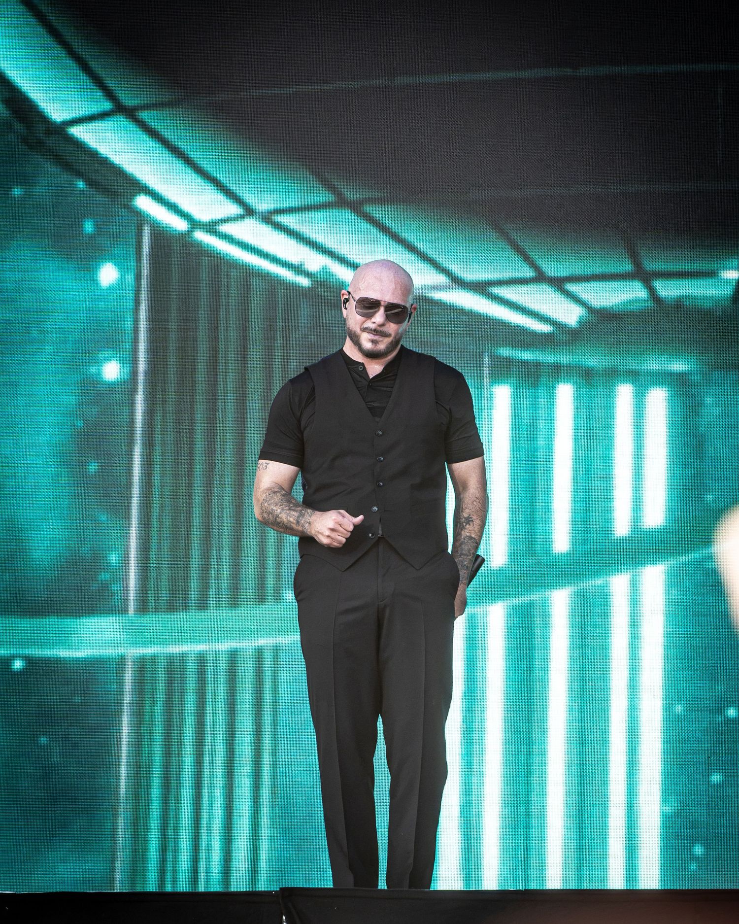 Bald man in black vest and pants smiling on stage with teal LED light backdrop at live performance.