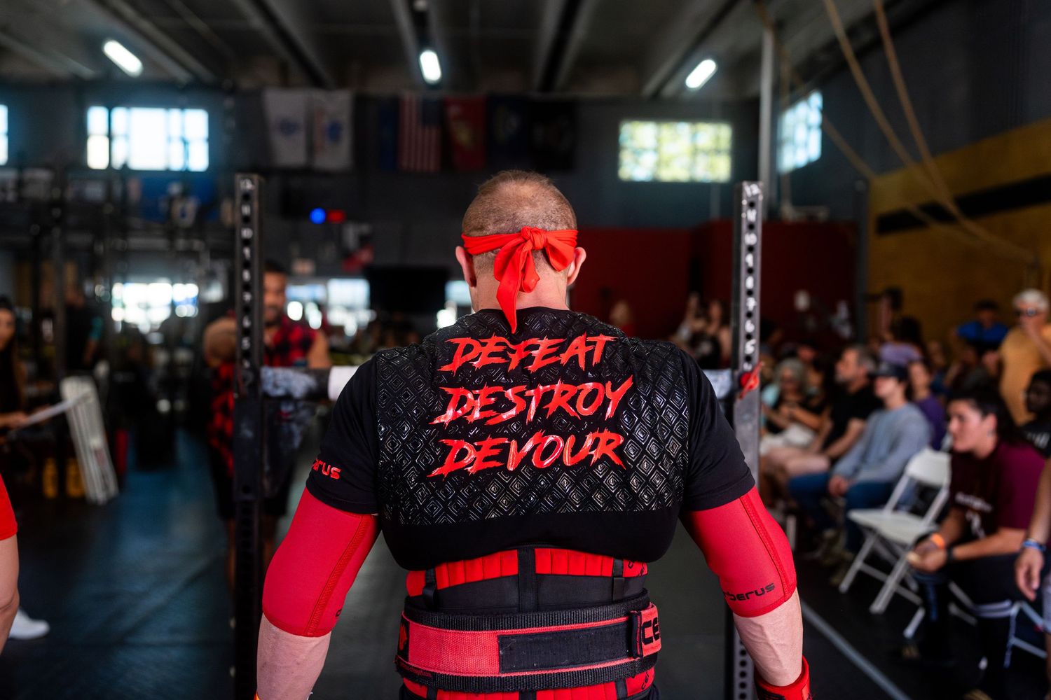 Athlete wearing red face mask and black 'Defeat Destroy Devour' t-shirt at indoor competition venue.