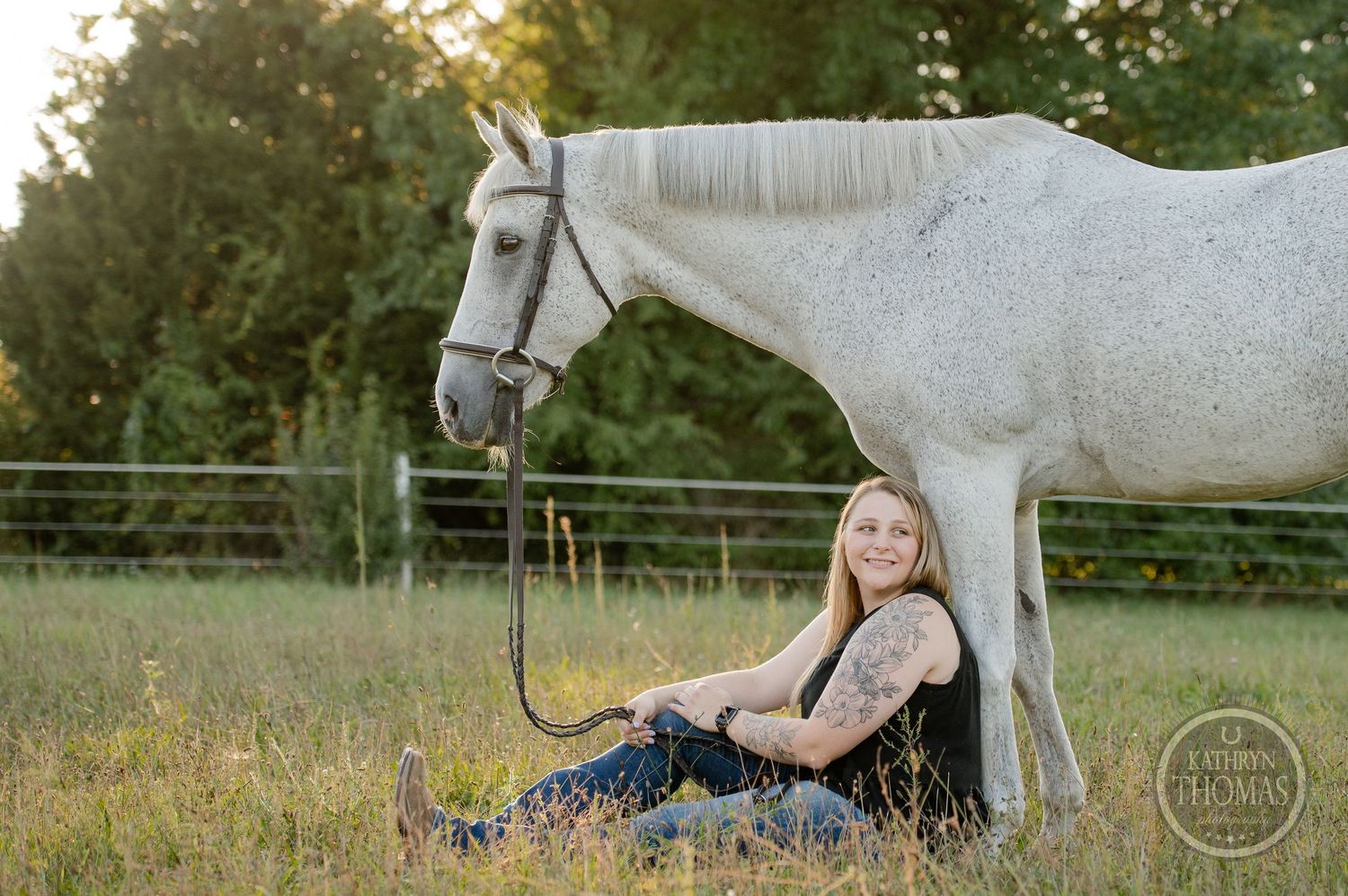 A person reclines on the ground beside a white horse in an outdoor setting.