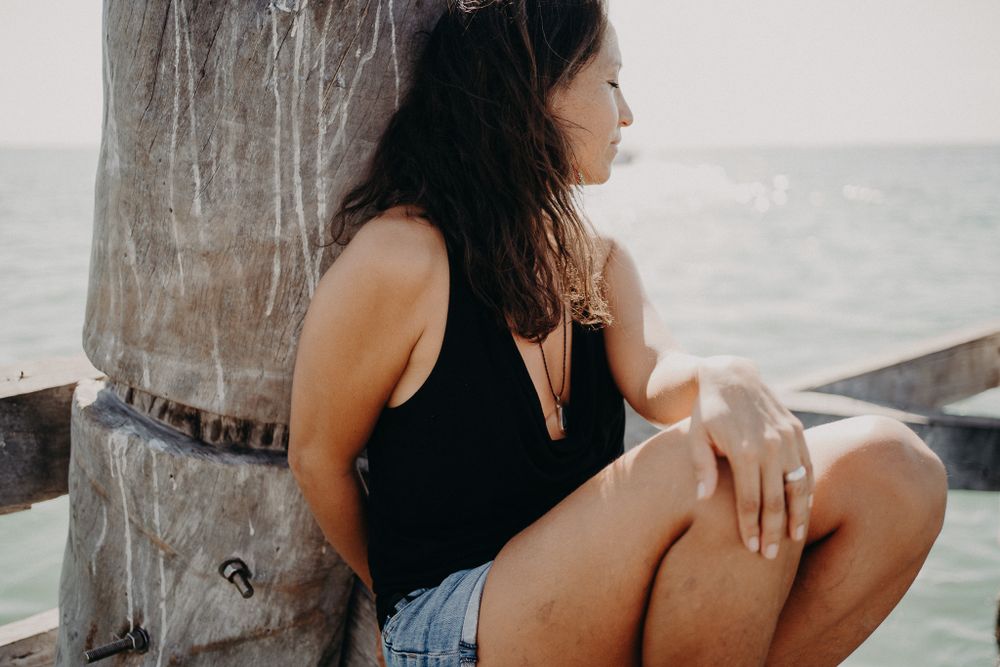 A woman in black tank top and denim shorts sits on a weathered pier by the ocean photoshoot in Puerto Morelos.