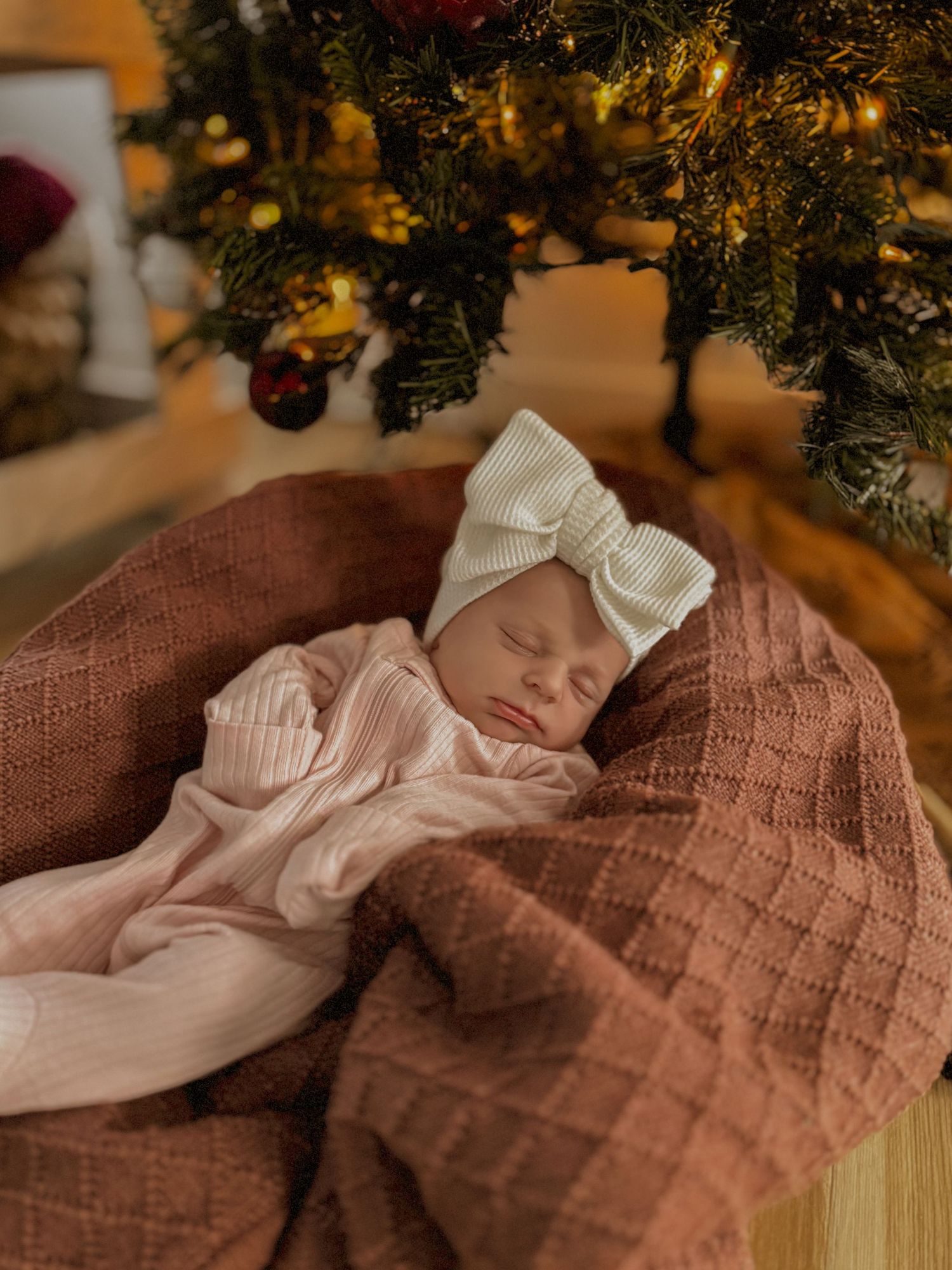A sleeping baby in white clothing rests on a brown blanket near a decorated Christmas tree.
