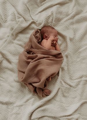 A sleeping newborn baby wrapped in brown fabric on white textured bedding.