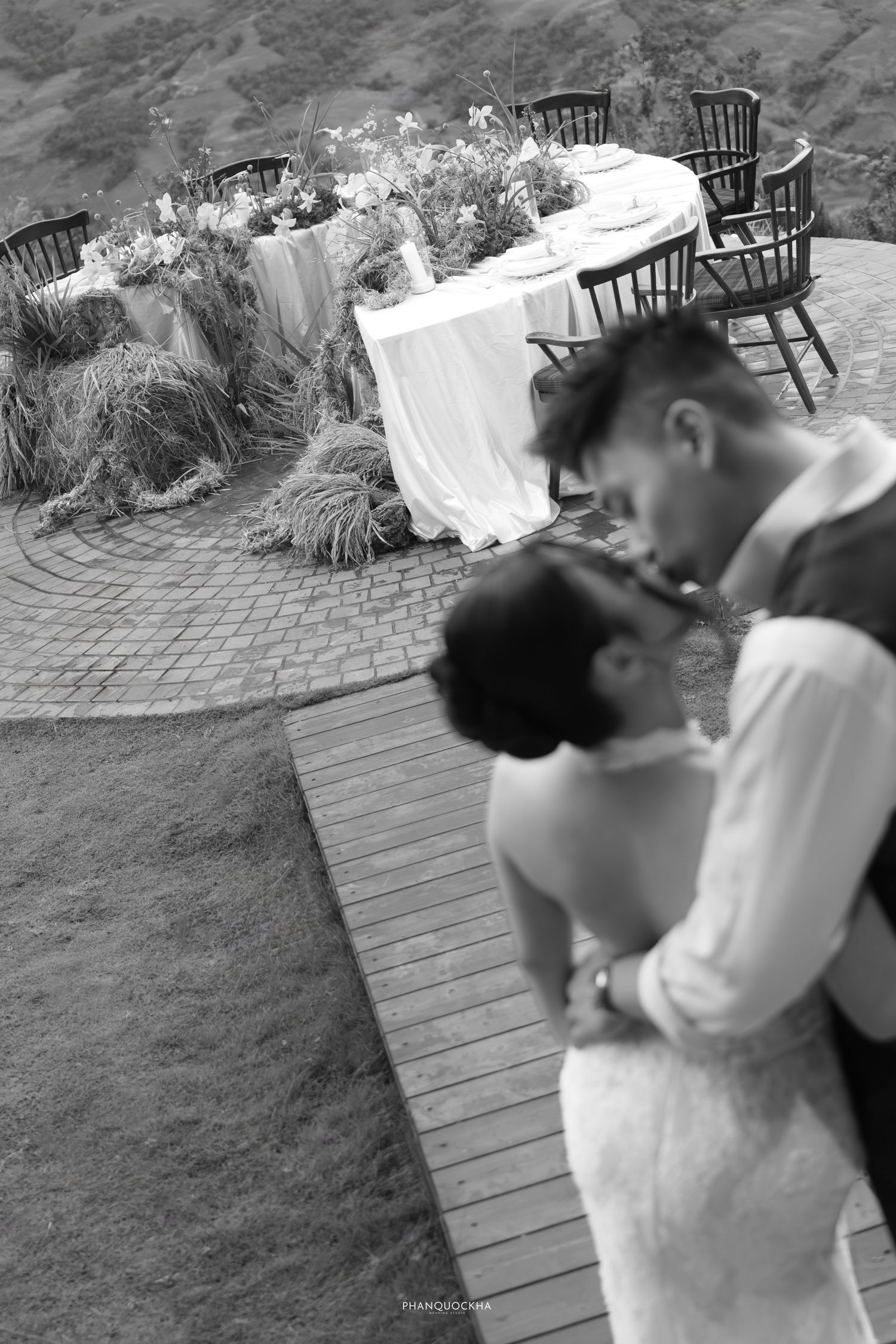 Black and white photo of bride and groom sharing a kiss near a decorated outdoor reception table on a mountain.