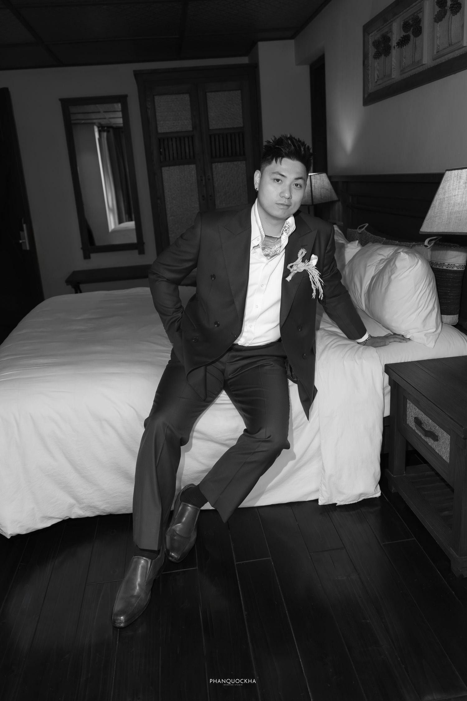 Black and white portrait of groom in dark suit sitting on a hotel bed with a boutonniere on his lapel.