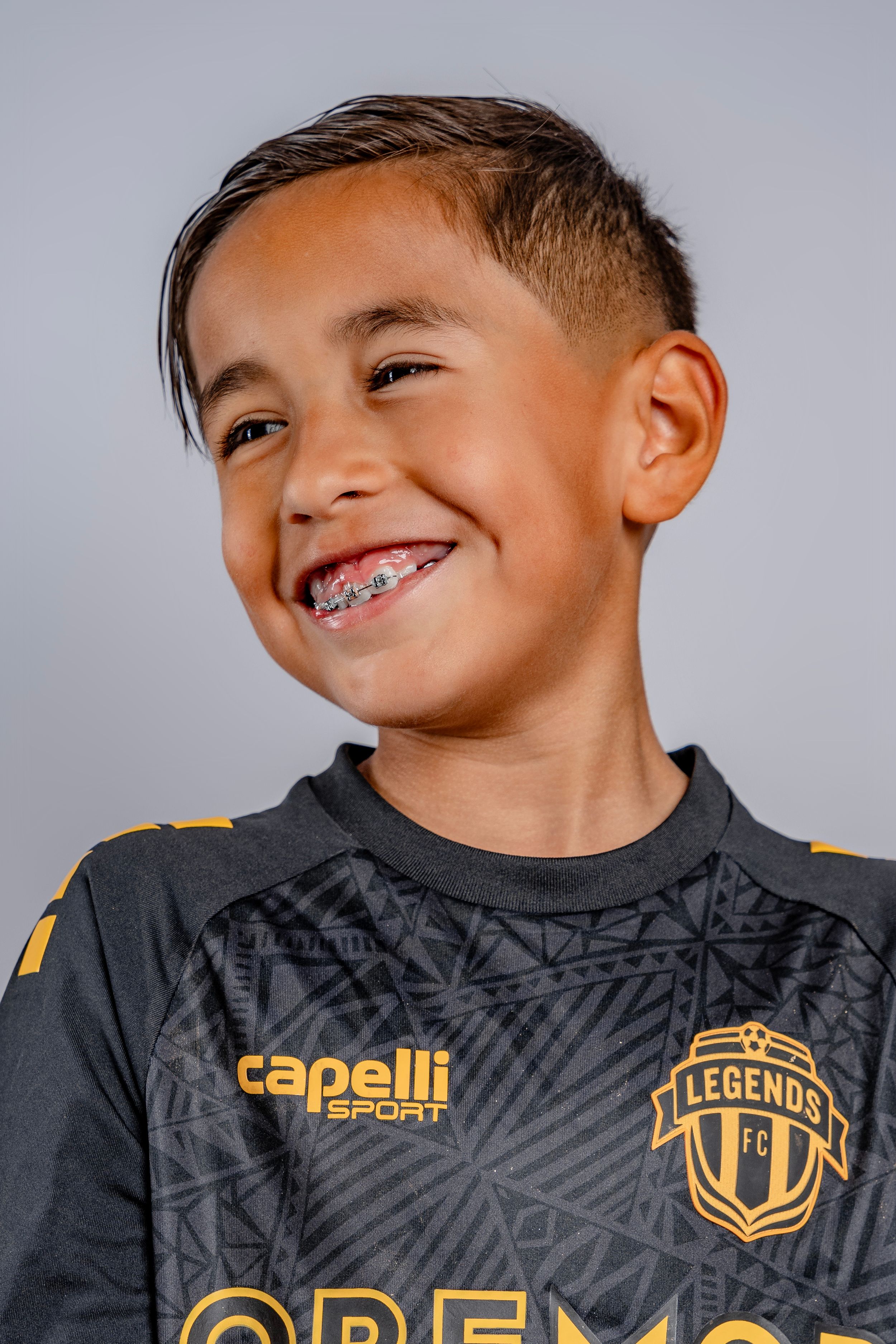 Young soccer player smiles playfully while wearing a black and gold Legends FC team jersey against gray background.