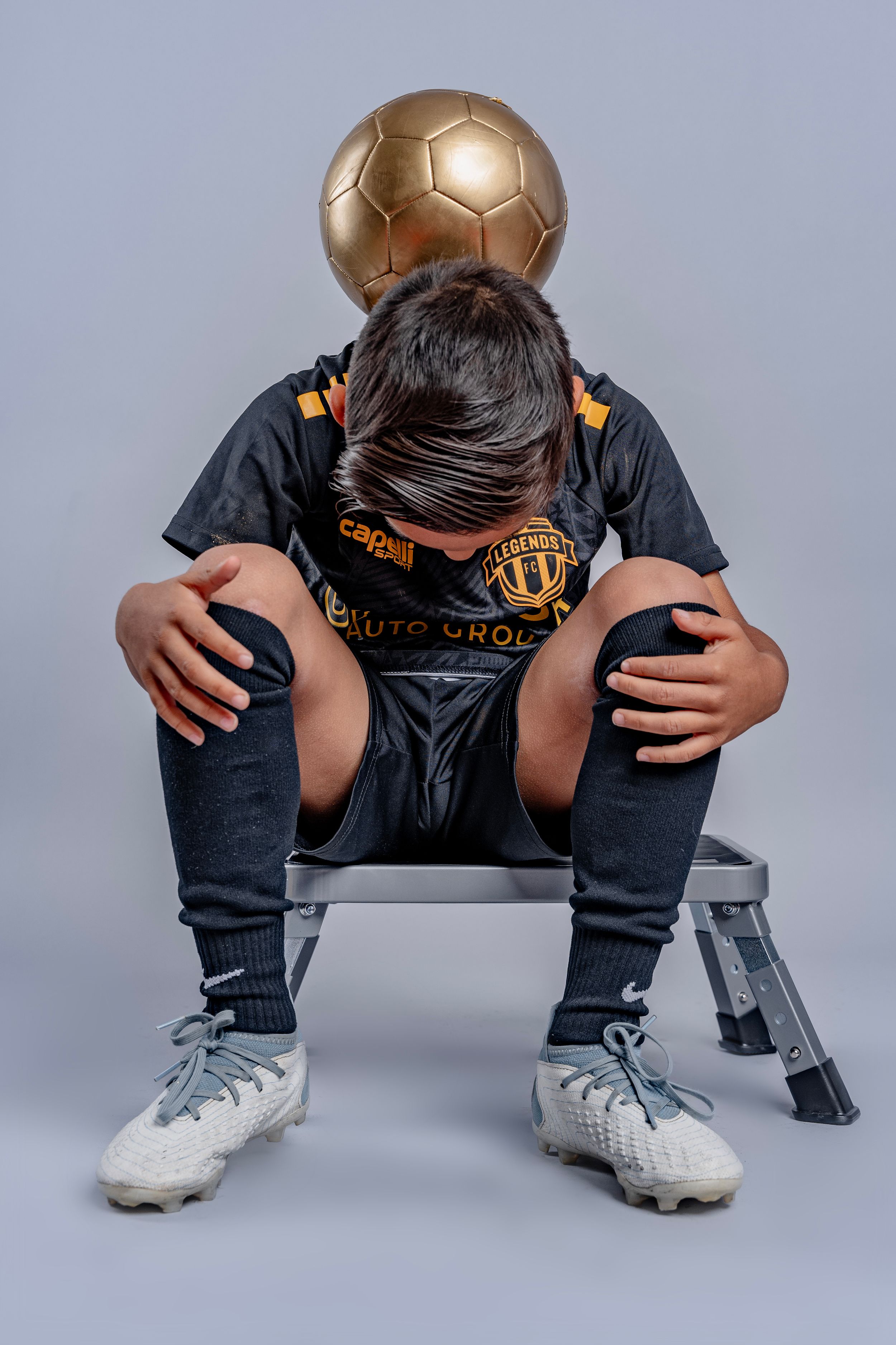 Young athlete crouches with gold soccer ball while wearing cleats and dark uniform.