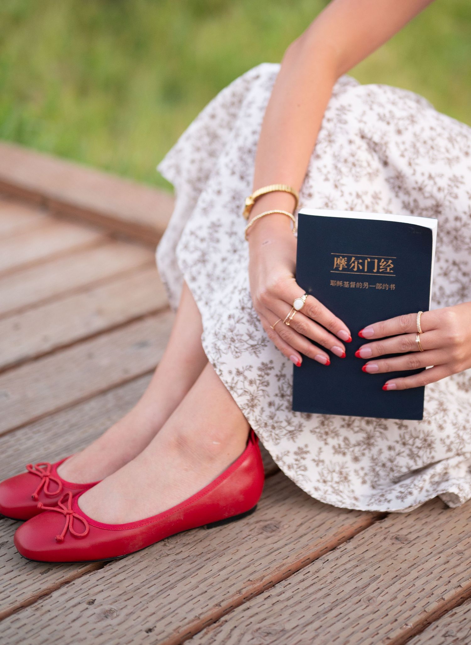 A person in a floral dress and red flats sits with a religious book on wooden decking.