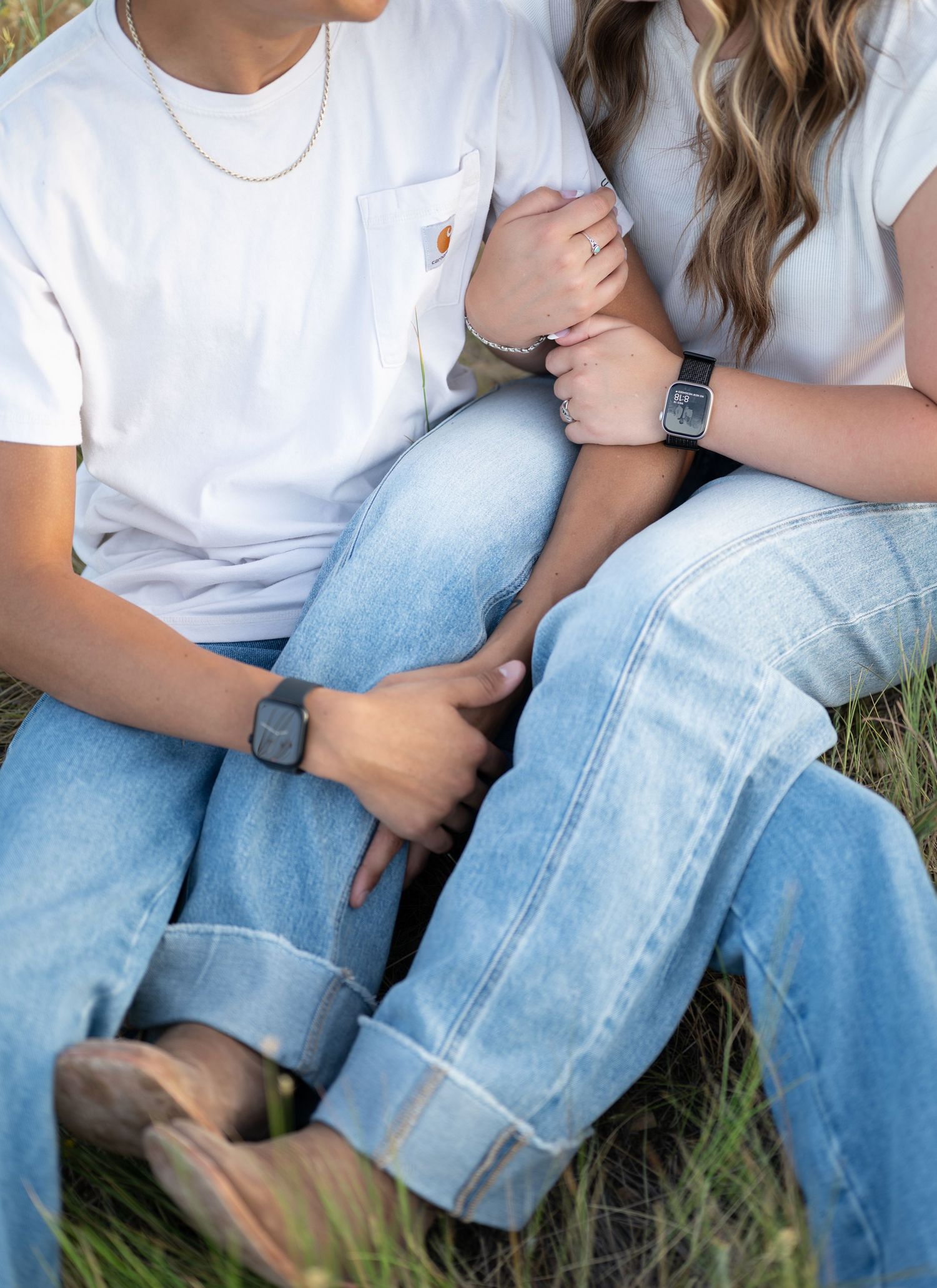 A casual couple in jeans and white t-shirts sit close together on grass.
