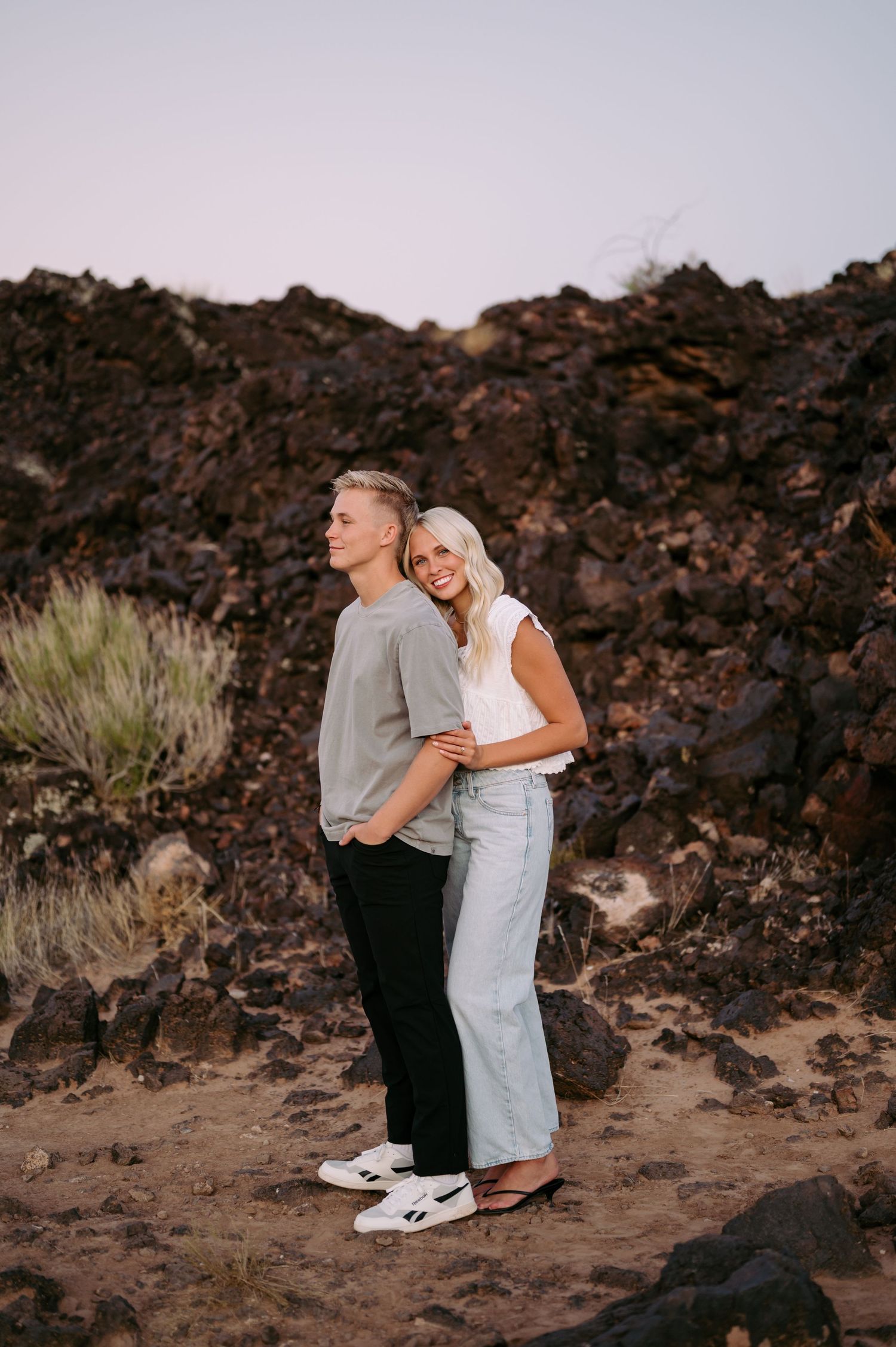 A couple embraces against a dramatic rocky volcanic landscape at sunset.