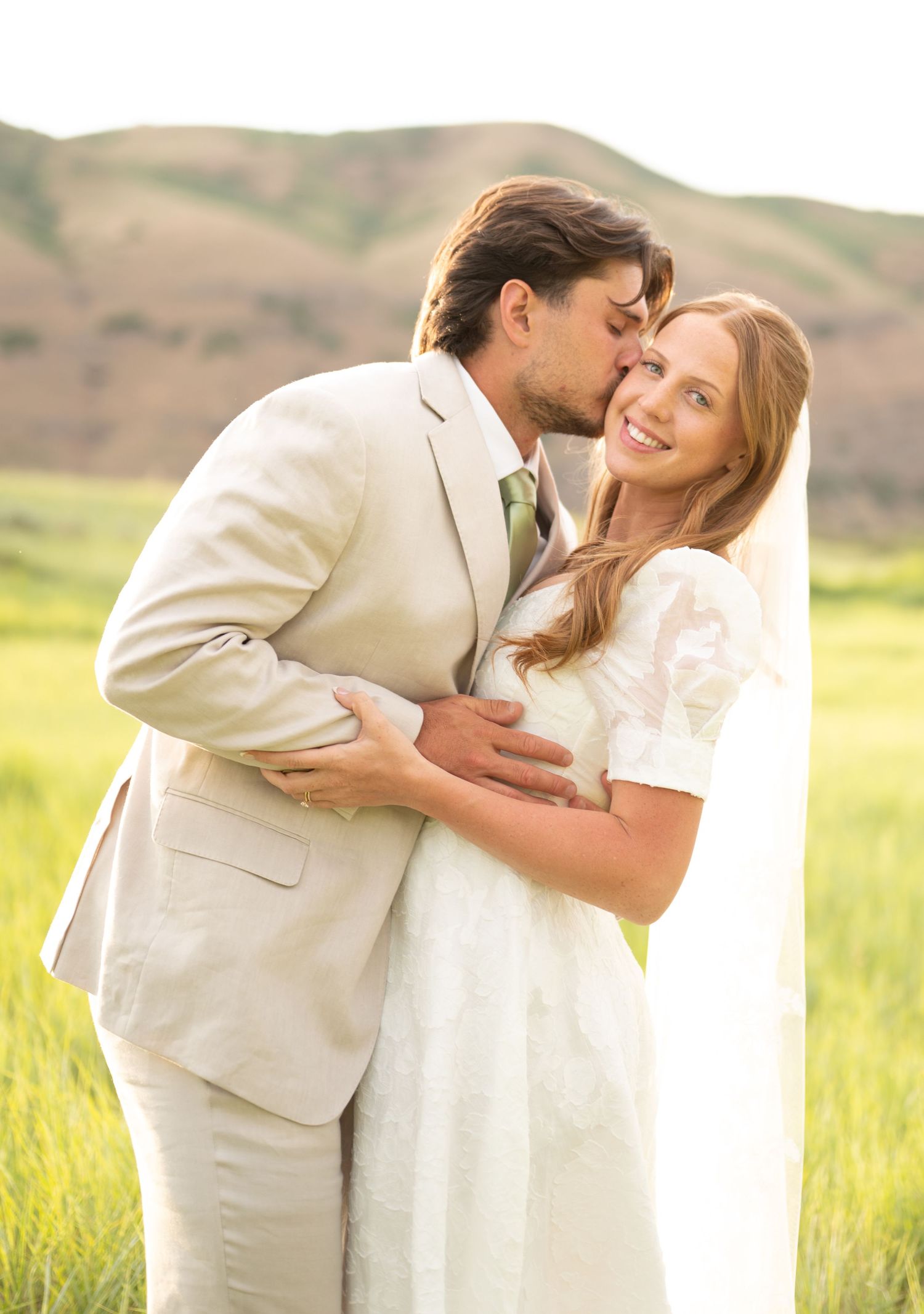 A couple shares an intimate moment in a sunlit field wearing light colored formal attire.